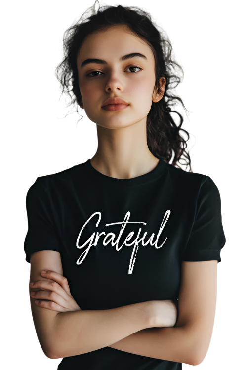 A young woman with dark, wavy hair pulled back in a loose, casual style stands against a neutral background. She has a calm and confident expression with her arms crossed. She wears a black T-shirt featuring the word "Grateful" in elegant white script across the chest. The minimalist design and contrast between the black fabric and white lettering make the message stand out clearly.