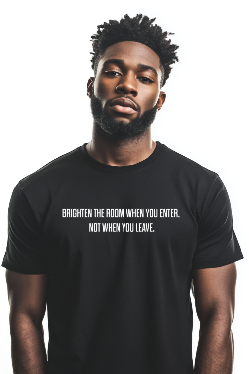 A young man with dark skin, a well-groomed beard, and short curly hair stands against a plain white background. He wears a black T-shirt with a motivational quote in white capital letters that reads, "BRIGHTEN THE ROOM WHEN YOU ENTER, NOT WHEN YOU LEAVE." His expression is calm and confident, and he has a relaxed posture. The T-shirt fits well, emphasizing its sleek and modern design.